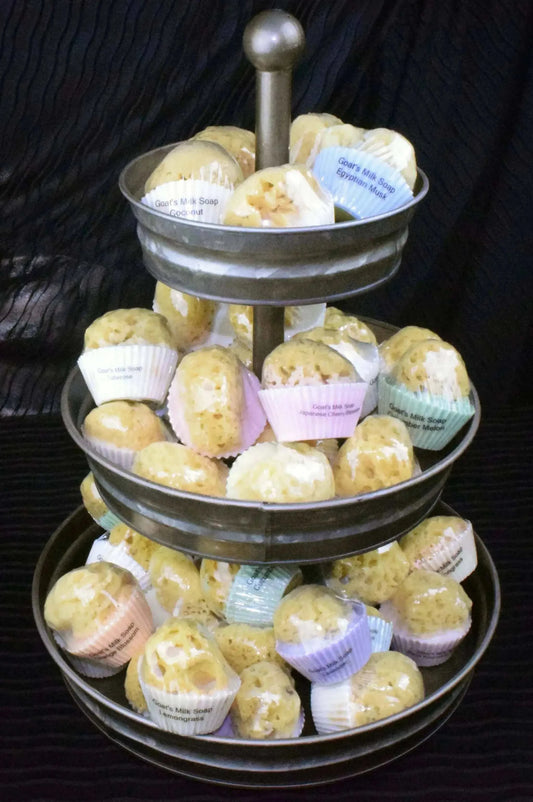 Three-tier galvanized steel display stand filled with goats milk and olive oil soaps shaped like cupcakes with natural sea sponges embedded, each soap in colorful paper cups.
