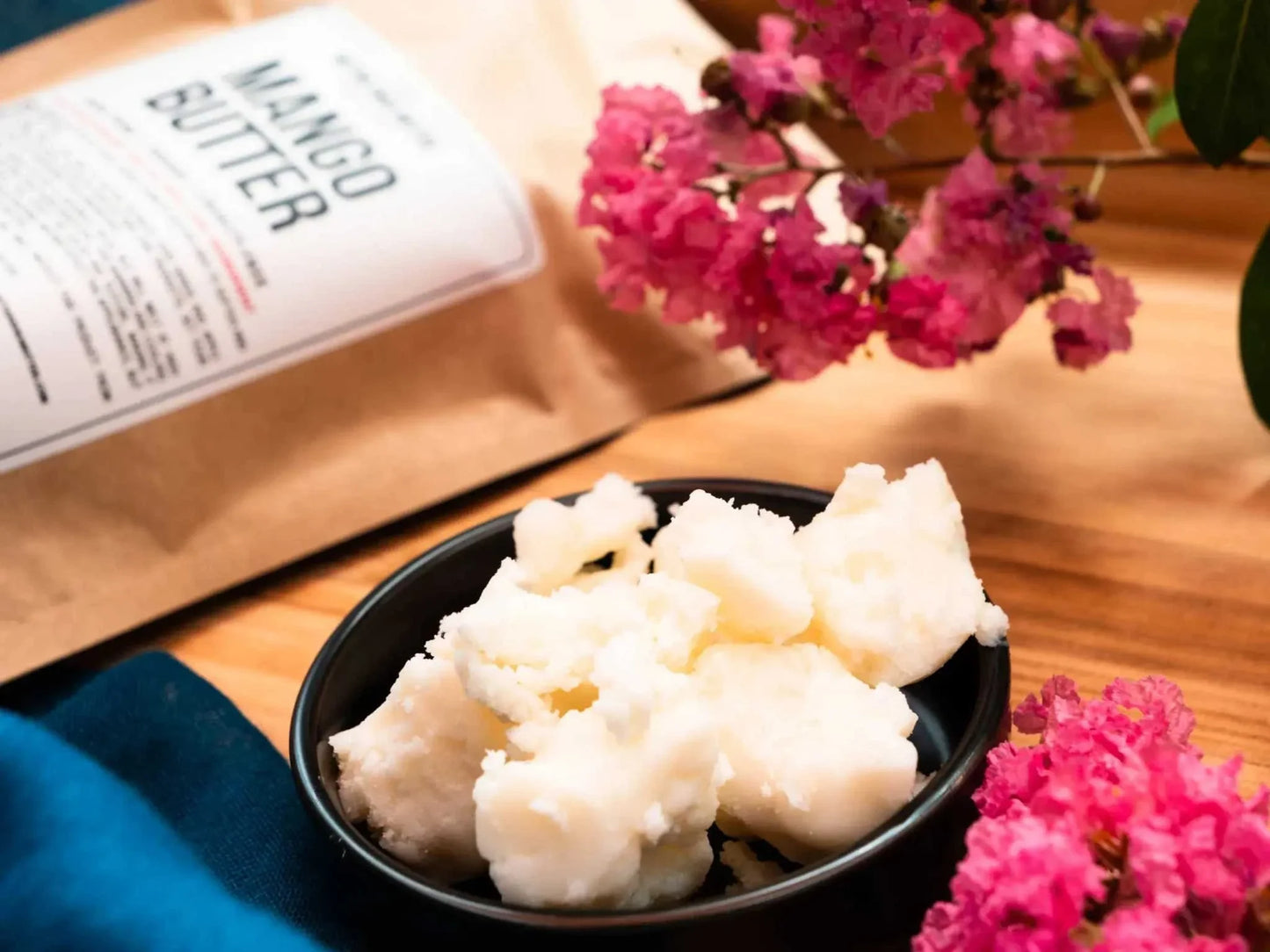 10lb blocks of pure mango butter bulk in a black bowl with packaging and pink flowers on wooden surface