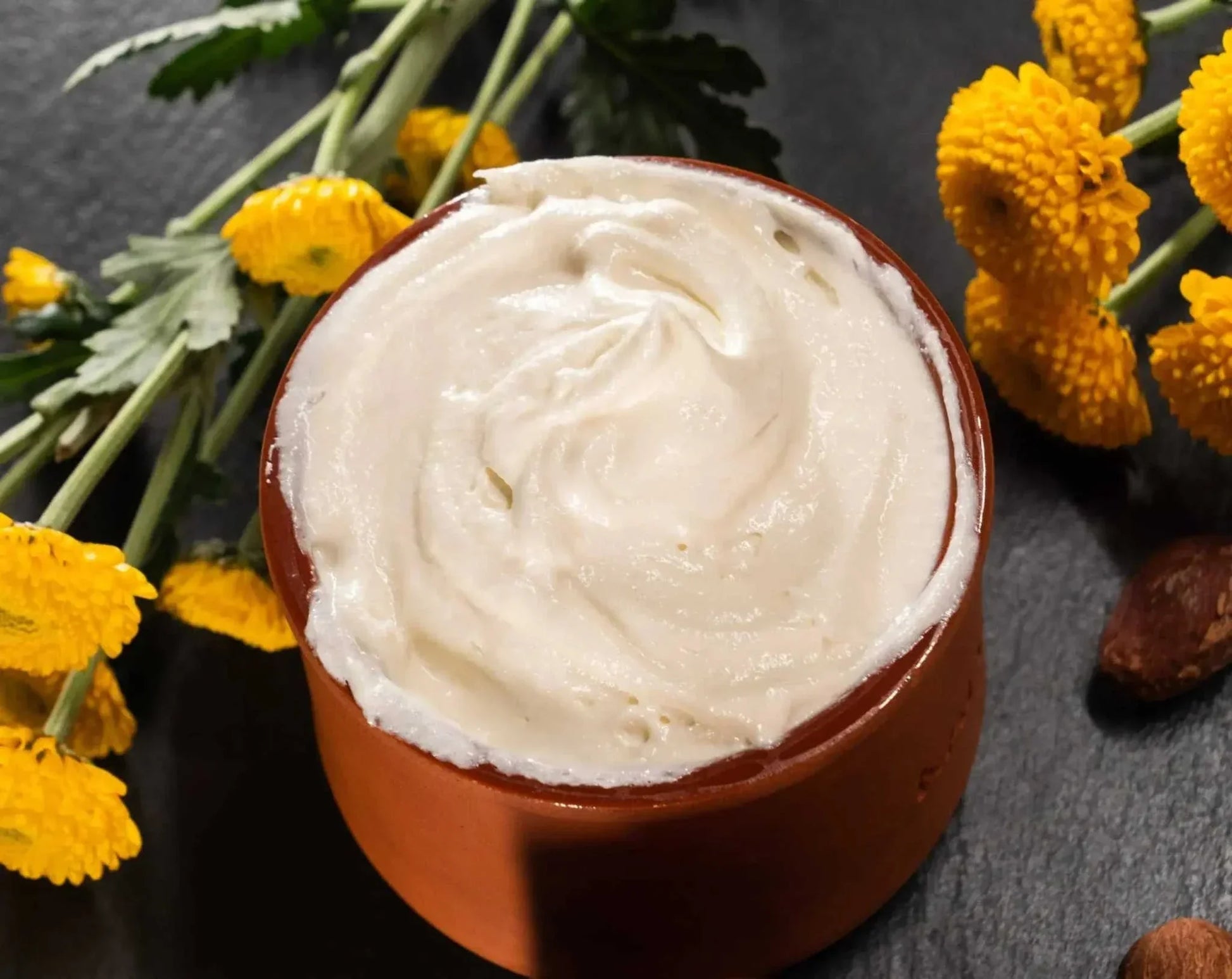 Bulk raw and unrefined shea butter in a brown bowl surrounded by yellow flowers for skincare and haircare use